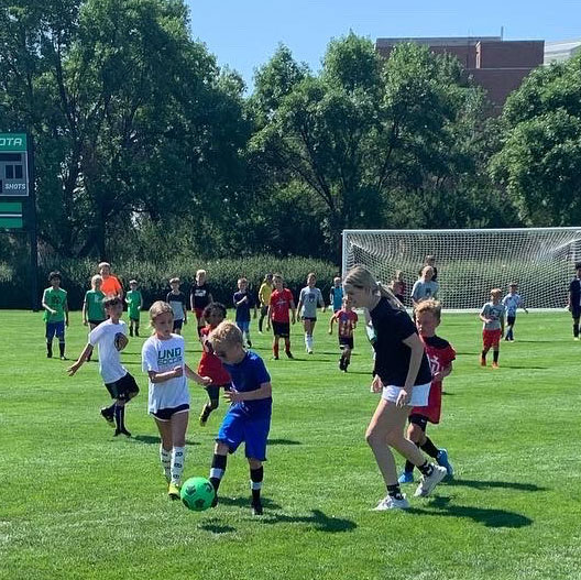 Kids-playing-soccer-at-UND-soccer-camp-in-Grand-Forks,-ND---sq Kids play soccer with coach on field at the University of North Dakota Soccer Camp in Grand Forks, ND
