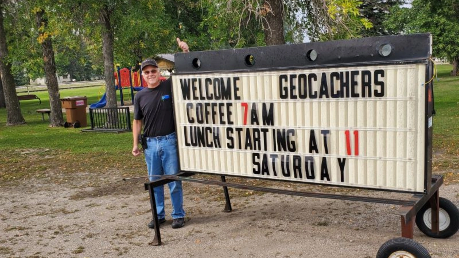 Geocaching-Grand-Forks-North-Dakota-Gilby-Welcome-Geocachers Geocaching in the Greater Grand Forks Region. Man stands in front of sign that reads "Welcome Geocachers."
