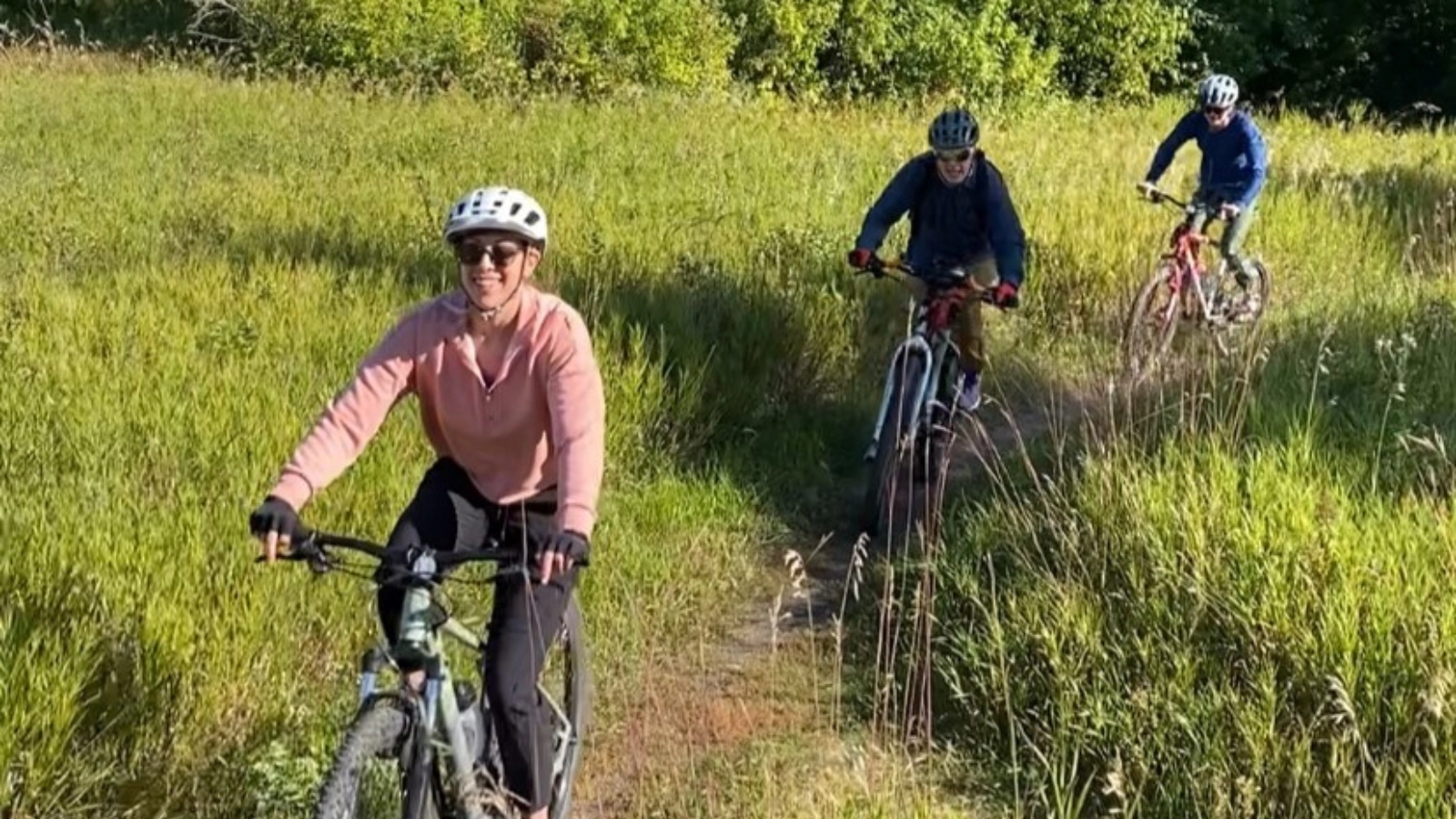Three people ride their bikes in a line on a worn down, grassy trail.
