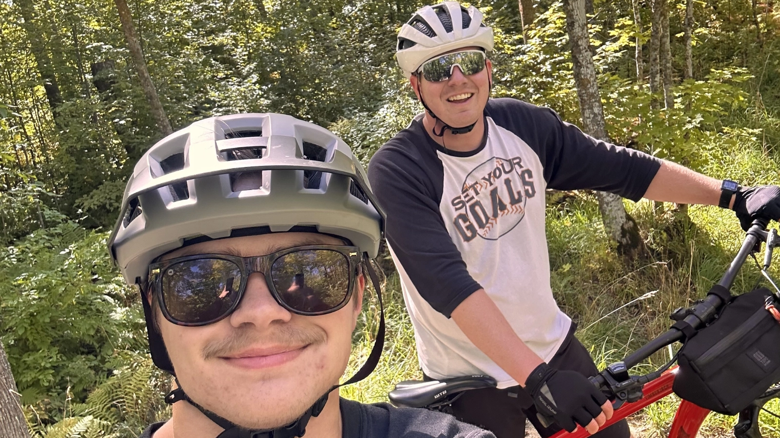 WCT writer Paul and a friend wear sunglasses and a helmet as they smile and bike through a heavily wooded area.