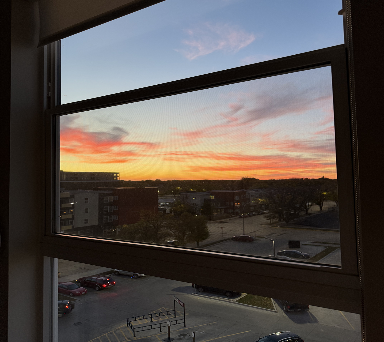 Sunset photo This image is through the window of an apartment building. It looks out onto a sunset from Grand Forks. The colors of orange, yellow, and pink bleed into the evening sky.