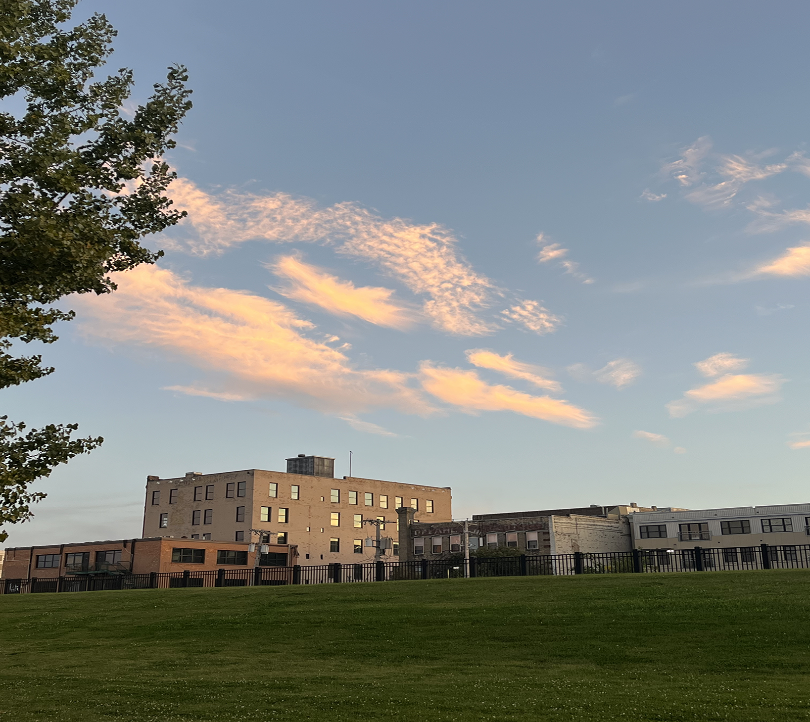 Building and Colorful Clouds photo This image looks at a green field with a building in the back of the image. The dusk sky takes up the majority of the shot with colorfully, orange and pink lit clouds floating through a blue sky above.