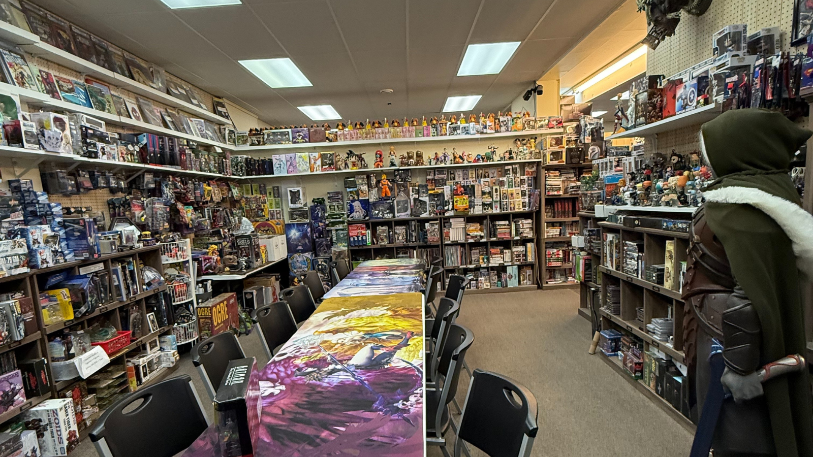 this image is from within the gaming store and displays dozens if not hundreds of games on the shelves. There is a long table going straight through the room that is decorated in gaming decor. Also an elvin mannequin stands to the right of the image holding a fake axe.