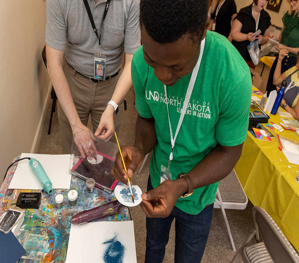A UND student is pictured participating in an art volunteering event through the InternGF Summer Cohort series. He wears a bright green UND shirt and his holding his art piece delicately while painting it.