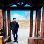 Author Ben stands under a wooden archway with a clear blue sky behind him as he smiles off to this distance.