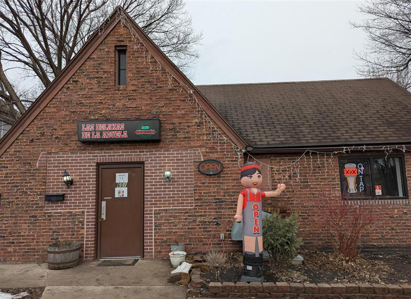 Outdoor photo of Las Delicias de la Abuela in Grand Forks. A small red and brown brick building with an statue of what looks like "Abuela" outside the front.