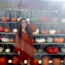 Natalie pumpkin wall photo Author Natalie poses in front of the Nelson Pumpkin Patch pumpkin wall in a burnt orange dress.