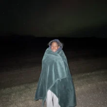 Precious Northern Lights Author Precious stands and smiles at the camera during a northern lights chasing trip while she's wrapped up in a cozy blanket!